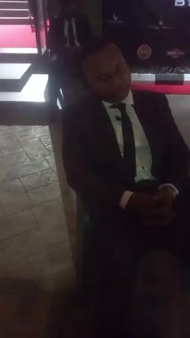 A medium shot shows a man in a black suit and tie sitting down. He has his eyes closed, and his hands are clasped in his lap. Behind him, a red carpet leads up to an entrance with a set of stairs. A person in a suit is standing near the entrance. A large sign with logos for liquor brands is also behind the seated man. The video is a bit shaky, and the lighting is subdued. The floor appears to be tiled.