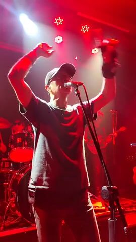 The video shows a man singing on stage, under bright red stage lights. He is wearing a grey t-shirt, dark pants, and a baseball cap. He has his arms raised in the air and then spreads them out. Behind him is a drum kit and a partial view of the rest of the band. The background is dark except for the stage lighting.