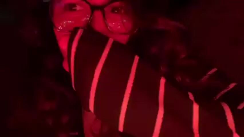 A person wearing glasses and a striped shirt is visible in a red-lit environment. Their face is partially obscured by their hand and the angle of the camera. The lighting is predominantly red, with some lights reflected in the glasses.