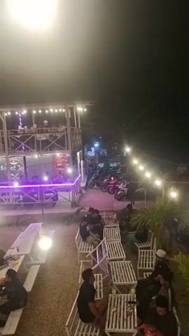 The video shows an outdoor restaurant at night. The restaurant has multiple levels and is decorated with string lights. People are seated at picnic tables and benches, eating and socializing. There are also motorcycles parked in the background. The atmosphere appears to be relaxed and casual.