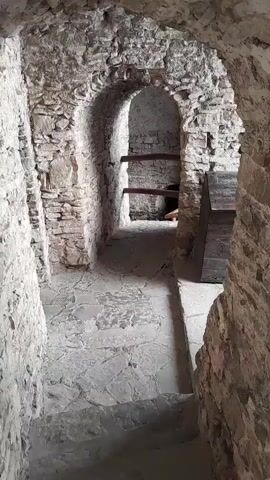 The video shows a stone passageway with arched doorways. The walls are made of rough-hewn stone, and the floor is paved with irregular flagstones. In the background, there are wooden beams and what appears to be a stone structure. The lighting is dim, suggesting an interior space like a cellar or an ancient building. There are no people visible in the video. The camera is positioned to look down a narrow corridor, with a set of stone steps leading downwards. The overall impression is of an old, possibly historical, underground area.