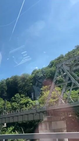 The video captures a scenic view from a moving vehicle, showcasing a lush, green hillside, a bridge, and a river under a clear blue sky. The scene is set in bright daylight, indicated by the strong sunlight and visible lens flare.

In the foreground, a metal guardrail suggests the viewpoint is from a road. Below the bridge, a river flows with visible white rapids, indicating a fast current. Above the river, a multi-sectioned concrete and metal bridge spans the landscape. This bridge appears to carry a road or railway, with supports and overhead lines visible along its length.

Behind the bridge, a steep, heavily forested hillside rises, covered in dense green trees and vegetation. The bright sun illuminates the top of the trees and casts a dappled light on the slope. The sky is largely clear, with a few wispy clouds. No people or specific activities other than the apparent travel are visible. The overall impression is one of a natural, mountainous or hilly landscape with significant infrastructure, typical of the Chvizhepse region in Russia.