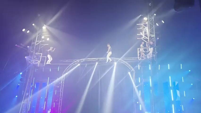 This video shows a performance at a venue with dramatic lighting. The main subjects are several individuals, likely performers, engaged in a high-wire act. Two individuals are prominently positioned on a thin wire stretched between tall metal structures. One performer stands with their arms outstretched, seemingly balancing, while another is also standing on the wire. Several other individuals are visible higher up on the metal scaffolding, some appearing to be part of the act or crew.

The scene is set within a dark indoor space, illuminated by bright stage lights that cast beams of white and blue light downwards and outwards. The lighting creates a dramatic and ethereal atmosphere. There are large, vertical panels with a patterned design, some illuminated in purple and blue, which suggest a stage or performance area. The overall impression is that of a circus or acrobatic show. The performers are dressed in white, form-fitting outfits, making them stand out against the dark backdrop and bright lights. The activity taking place is a skilled acrobatic performance, likely involving balancing and coordination on the high wire. There are no visible indications of the time of day, weather, or specific location beyond the performance setting itself. The mood is one of awe and excitement, typical of a live performance.