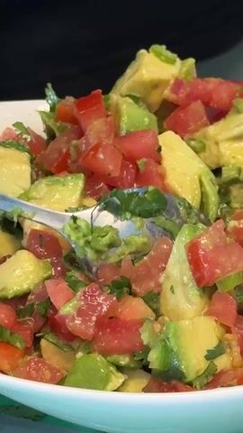 A close-up video shows a white bowl filled with a chopped salad. The salad's ingredients are primarily red tomatoes and green avocado, both cut into small, roughly equal-sized cubes. Some green herbs, possibly cilantro, are also visible, mixed throughout. A silver spoon is used to stir the salad, gently mixing the ingredients. The background is dark and out of focus.