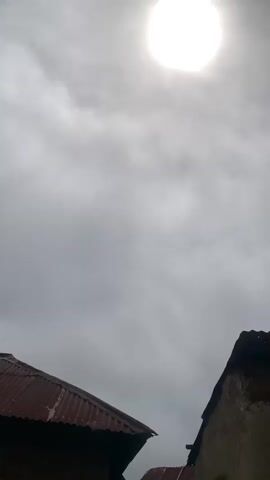 The video shows a daytime outdoor scene. In the foreground, two structures with corrugated metal roofs are visible. The one on the left is darker and closer to the camera, while the one on the right is lighter and further away. The sky above is overcast with a bright sun visible through the clouds. Several people are partially visible in the lower part of the frame. On the left, a person in a yellow shirt and a person wearing a head covering are seated. In the center, a person in a blue hooded sweatshirt is also seated. On the right, a person wearing a white head covering is standing. There are also some large pots or containers visible on the ground between the structures. The ground appears to be dirt.