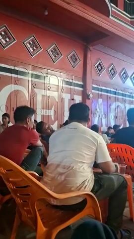 This is a video of people sitting and watching a projected image of a sporting event. The projection screen is large and bright, showing a field with players. The audience is seated on orange plastic chairs, facing the screen. The environment appears to be an outdoor seating area, possibly a restaurant or cafe, with red-painted walls and decorative elements visible in the background. The lighting suggests it is nighttime. Several individuals are visible from behind, suggesting a communal viewing experience.
