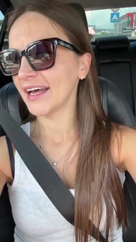 A young woman with long brown hair and sunglasses is sitting in the driver's seat of a car. She is wearing a white tank top and a seatbelt. The car interior is visible, including the car seat and the seatbelt. Outside the car, a road with traffic signs and buildings can be seen. The woman appears to be looking at the camera.