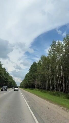 The video shows a view from a moving vehicle on a highway. The road has two lanes in the direction of travel, with a white line dividing them. On the right side of the road, there is a dirt shoulder and a dense forest of tall green trees. On the left side of the road, there is also a line of trees. The sky is partly cloudy with patches of blue sky visible. Two white cars are visible on the road ahead, traveling in the same direction as the viewer's vehicle. The lighting suggests it is daytime.