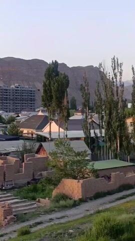 The video shows an aerial view of a semi-rural area with mountains in the background. In the foreground, there is a partially constructed building made of bricks, and other low-rise houses and buildings. In the mid-ground and background, there are more houses, some with green roofs, and several multi-story buildings under construction, one of which is covered in green netting. Two large yellow cranes are visible, indicating ongoing construction. The sky is a pale, hazy orange, suggesting either sunrise or sunset. The overall impression is of a developing community situated in a mountainous landscape.