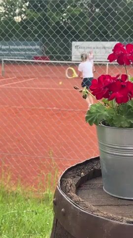 A blurry shot shows a young female tennis player in a white t-shirt and blue skirt hitting a yellow tennis ball on a clay court. She is facing away from the camera and is in mid-swing. The court is enclosed by a wire fence. In the foreground, a metal bucket with red flowers and green leaves sits on a wooden barrel. Another person can be seen in the background, also on the court. The scene appears to be outdoors during the day.