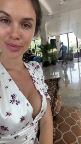 The video opens with a close-up selfie of a woman. She has dark hair pulled back, fair skin with some visible blemishes, and full, pink lips. She's wearing a white dress or robe adorned with a pattern of pink and red roses and green leaves, featuring a deep V-neckline that reveals cleavage. She offers a subtle smile, and her eyes are initially slightly closed before opening. Next to her, a cushion with a leopard print pattern is visible.

As the video progresses, the camera slowly pans to the right, revealing the expansive interior of what appears to be a large, elegant hall or hotel lobby. The space features a high white ceiling with decorative moldings and recessed lighting. Large white pillars support the structure, and the floor is made of highly polished light-colored tiles, reflecting the ceiling and windows.

In the mid-ground, a man in a dark t-shirt and dark pants can be seen walking from left to right, carrying a blue bag. Several large potted palm plants are placed throughout the area, adding greenery. Through large windows in the background, natural light streams in, and glimpses of trees or outdoor foliage are visible. Various pieces of equipment, including speakers and stands, are set up, suggesting preparations for an event or a post-event cleanup.

Further into the pan, another individual is seen in the distance, near a large white draped curtain or screen, bending down and appearing to be engaged in work. The general atmosphere suggests activity, likely the setting up or dismantling of an event in this grand, well-lit venue. The patterned cushion next to the woman, now more clearly a brown and white geometric or animal print, remains in the foreground.