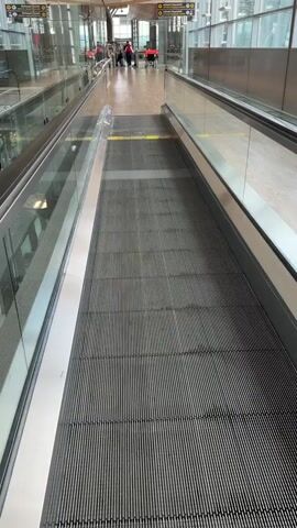 The video shows a moving walkway or travelator in a modern airport terminal. The walkway is empty and moving downwards, away from the camera. The walkway has a textured grey surface with parallel lines. On either side of the walkway are glass partitions with metal railings, separating it from the surrounding areas. In the background, there are signs indicating gate information. A few people are visible in the distance, some with luggage and red wheelchairs, suggesting they are passengers. The lighting in the terminal is bright and airy, with a high ceiling and large windows. The overall environment appears clean and organized.