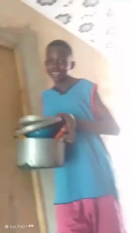 A young person, possibly a child or adolescent, stands in front of a wooden door and a light-colored wall. They are smiling and holding a large metal pot with a lid. The person is wearing a bright blue sleeveless tunic over pink shorts. The pot has a dark, stained bottom, suggesting it has been used for cooking. The ceiling above is decorated with a repeating pattern of stylized gray flowers. The overall lighting suggests it is daytime, possibly indoors or in a covered outdoor space. A padlock is visible on the door. The scene appears to be in a residential setting. The text "© itel P40+" is visible in the bottom left corner, indicating the device used to capture the image. The location context is Achara Ndiagu Omegio, Nigeria. The person's expression is cheerful and engaging.