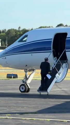 A man in a dark suit and light-colored shirt walks up a portable staircase attached to the open door of a white and blue private jet parked on an airport tarmac. The man is facing away from the camera and is ascending the stairs into the aircraft. The sun is shining and the sky is clear.