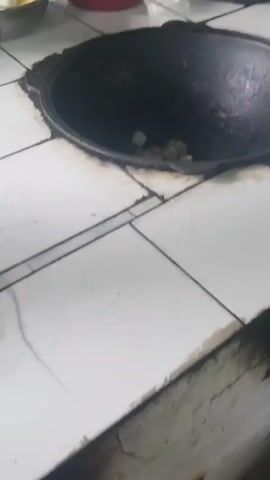 The video pans over a cooking scene. In the foreground, a large black cast-iron pot is situated in a tiled surface, with small white pieces of food in its bottom. To the left of the pot, there are two bowls on the tiled surface. The left bowl contains chopped potatoes and onions in a clear liquid. The right bowl holds chopped green peppers and red tomatoes. Behind the bowls, a man wearing a dark blue shirt and dark pants is visible. He is holding a patterned towel in his left hand and a utensil in his right hand. He appears to be preparing to add ingredients to the pot. The background shows a rustic outdoor setting with some wooden structures and greenery. The lighting suggests it is daytime.