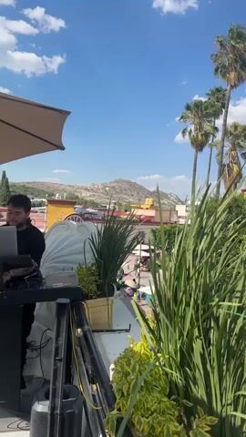 The video shows a town square on a sunny day, with palm trees, green bushes and small potted plants. In the background are mountains and a bright blue sky with fluffy white clouds. The camera pans over to show a church tower, then a peach-colored building with a clock tower on top. There are people milling about on the street in front of the buildings. Loud music is playing in the background.