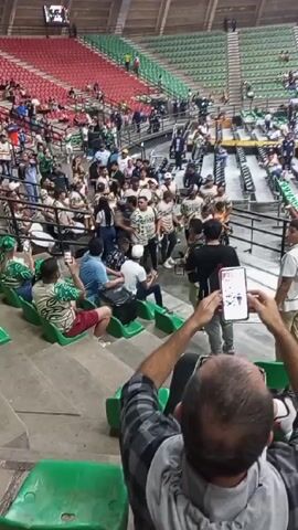 The video shows a group of musicians performing in a stadium. They're dressed in matching outfits, and the camera appears to be held by someone sitting in the stands, who is filming the performance with their phone. Other people in the surrounding seats are also holding up their phones to record. The stadium has green, red, and gray seating sections, and there are spectators scattered throughout.