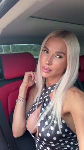 A woman is seated in the back of a car, directly facing the camera for a selfie-style video. She has long, straight, blonde hair and is wearing a black halter-neck dress with white polka dots, featuring a deep V-neckline. She has visible makeup, including lip gloss, and her expression is calm and direct. Her left hand is raised towards her face, near her ear, and she is wearing multiple bracelets on her left wrist and rings on her fingers. The car's interior features red and black seats, and a sunroof is visible above. Through the car windows, a blurred background of green foliage and trees can be seen, suggesting an outdoor, possibly rural or suburban, setting during the daytime. She poses and maintains eye contact with the camera throughout the short video.