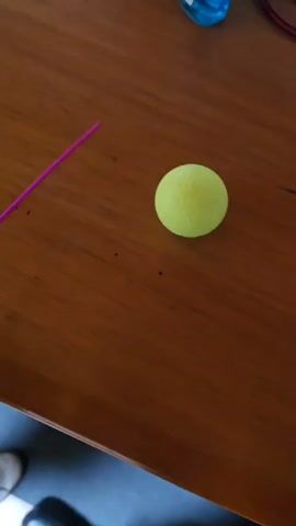 A hand with blue fingernails holds a pink stick with a yellow disc attached, which is then used to push a teddy bear wearing a navy blue uniform with gold stripes on its sleeves and a matching hat. The bear falls over on a brown wooden surface.