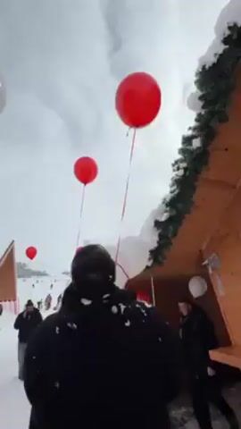 The video shows a snowy outdoor scene with red and white balloons. Several people are present, some in black jackets. A wooden structure with snow on its roof and evergreen decorations is visible. People are standing and walking around. The camera angle looks up towards the sky and balloons.