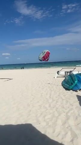 The video shows a beach scene on a sunny day. The sky is blue with scattered white clouds. A large, white and red parasail is suspended in the air over the ocean, seemingly pulling a person or persons. On the beach, a few individuals are standing near the water's edge. In the foreground, there are some beach chairs and turquoise-colored equipment lying on the sand. The sand is white and smooth, with a shadow cast in the lower left corner of the frame. The water is a mix of turquoise and darker blue.