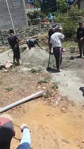 A group of men are working on a construction site. There is a large pile of sand and other building materials. Several men are shoveling sand, mixing it with cement, and transporting it. One man is using a shovel to mix the materials. Another man is standing and observing. There are tools and construction equipment scattered around the site. The scene appears to be outdoors during the day, with a sunny sky and trees visible in the background. The ground is dirt and gravel.