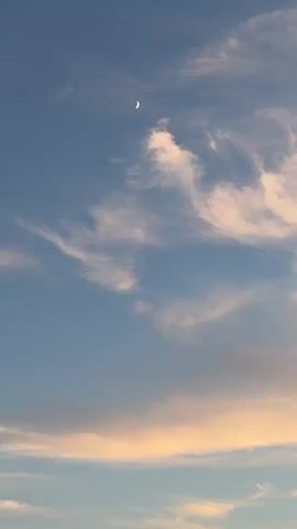 The video displays a soft blue sky with scattered clouds. A crescent moon is visible in the upper portion of the frame. The lower part of the sky is tinged with a warm orange hue, suggesting either sunset or sunrise. The clouds are wispy and ethereal, with some appearing brighter and more defined than others. The overall scene is serene and natural, with no discernible human presence or activity.