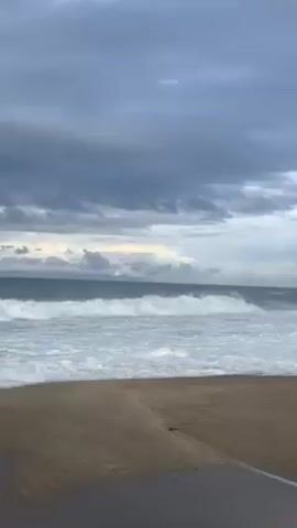 The video shows an overcast sky above a beach and ocean. Large waves are rolling in towards the shore. Later in the video, the camera pans to show two men standing on a concrete structure near the water’s edge, apparently fishing. There is plastic debris and a stool on the ground near them.