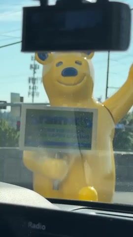 This is a first-person perspective video, likely from inside a car. The camera is angled upwards towards the windshield. Directly in front of the camera, and seemingly very close, is a large, bright yellow bear statue. The bear has blue accents on its ears and nose and appears to be smiling. It is holding a rectangular sign in front of its chest that displays the brand name "Rain-X" and text advertising "BETTER VISIBILITY MEANS SAFER DRIVING." The statue is wearing a thin black collar or strap. Behind the bear, the background suggests an outdoor, possibly urban environment with utility poles and some greenery. The lighting indicates it is daytime with a clear sky. In the foreground, a black dashboard with a screen displaying "FM Radio" and "107.7" is partially visible. The video has a shaky quality, suggesting movement.