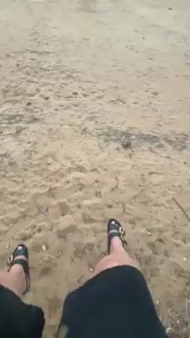 A first-person perspective video shows an individual sitting on a sandy dune or beach with the ocean in the background. The person is wearing black shorts and black sandals. The sand is a light tan color. The ocean is visible on the horizon with a cloudy sky above. The camera pans slightly to show more of the beach and the ocean view. There are some bushes and a light post on the horizon near the water.