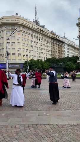 The video shows a group of people performing a traditional dance in a plaza, surrounded by buildings. Several couples dance in a circle. The dancers wear traditional Argentine clothing, including long skirts, capes, hats, and black pants. The plaza is paved with cobblestones. The sky is overcast. Some people are sitting on benches in the background, watching the dancers. A police vehicle is parked in the background. Billboards are visible on buildings.