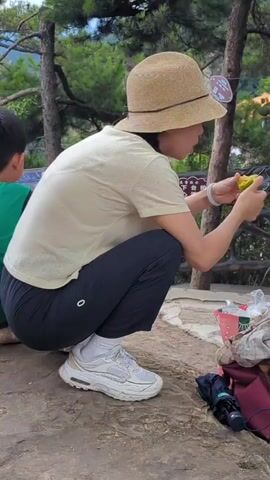 A woman wearing a straw hat, a light-colored t-shirt, dark pants, white socks, and white sneakers is squatting on a rock. She is holding a piece of fruit in her right hand and appears to be eating it. To her left, a portion of a person wearing a green shirt is visible. In the background, trees and foliage are present. To the woman's right, a brown bag, a red bag, and a small white container with items inside are on the ground.