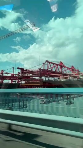 The video shows a construction site with a large red crane and other construction equipment. The crane is visible against a blue sky with white clouds. A bridge or similar structure is under construction, with its framework visible. The camera appears to be moving, possibly from a vehicle, as evidenced by the blur and the presence of a barrier in the foreground. The lighting suggests it is daytime. There are no people clearly visible in the frame. The overall scene depicts an active construction project.