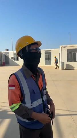 The video shows a person in a yellow hard hat and an orange safety vest with a blue reflective vest over it, standing outdoors. The person is wearing a black face covering and sunglasses. They are holding a black object, possibly a walkie-talkie, in their right hand. In the background, there are several white modular buildings and another person wearing a hard hat and safety vest walking away from the camera. The ground is paved and the sky is clear and blue. The person in the foreground is looking up and to the right. The video is shot from behind and slightly to the side of the main subject.