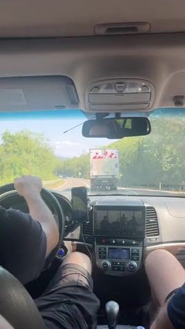 The video shows the interior of a car, looking forward through the windshield. A person's hands are visible on the steering wheel. The car is driving on a road bordered by trees. A truck is visible ahead, with what appear to be red hearts painted on the back. The car's dashboard screen displays a video of people sitting at a table. The overall impression is that of a road trip.