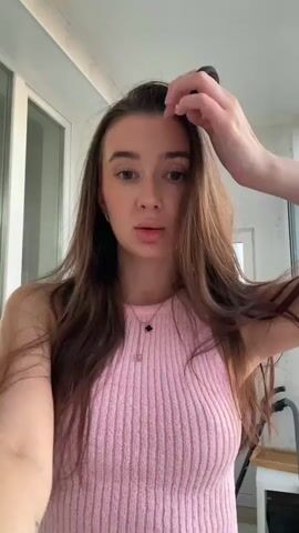 A young woman with long, brown hair and brown eyes is standing in front of a camera. She is wearing a pink, ribbed, sleeveless top and gray sweatpants. She is touching her hair with her hands. The background appears to be an indoor setting with a window and a door. There is also a chair and some kind of cleaning equipment visible. The lighting suggests it is daytime.