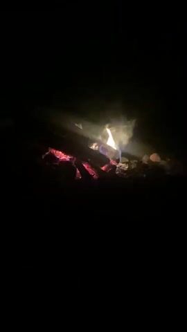 A close-up shot of a campfire at night. The fire is burning with visible flames, embers, and smoke. The background is completely dark, indicating it is nighttime. The camera is positioned low, close to the ground, focusing on the fire itself.