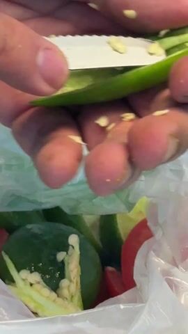 The video shows a person removing seeds from a halved green chili pepper with a paring knife. The seeds are removed over a clear plastic bag filled with additional halved chili peppers, limes, and red bell pepper. The pepper is held in one hand while the knife scrapes out the seeds. Some seeds stick to the person's fingers as they are removed. The background includes a white countertop.