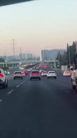 A first-person view from a vehicle on a multi-lane highway at dusk shows a steady stream of cars moving forward. The sky is a light orange and pink. Power lines and transmission towers line the left side of the road, and buildings are visible in the distance. The cars have their headlights and taillights on, and many appear to be silver or white.