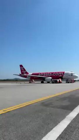 The video showcases an airport tarmac on a clear, sunny day. Initially, an AirAsia aircraft, painted in red with white lettering, is stationary on the tarmac. Then, a yellow Scoot plane appears in the background as passengers disembark from a white airplane via mobile stairs, which are staffed by an airport worker. People are walking across the tarmac from the mobile stairs. The footage includes the surrounding environment, such as trees and buildings in the distance.