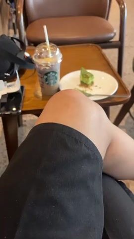 The video shows a first-person perspective of someone sitting at a table in a cafe. In the foreground, the person's knee and upper thigh are visible, clad in black shorts. On the table, there is a clear plastic cup with a brown beverage and whipped cream, bearing the Starbucks logo, and a white plate with a slice of green cake. A fork rests on the plate. To the left of the cup and plate is a black bag and a smartphone. In the background, other cafe patrons are seated at tables and chairs. A brown leather chair is directly behind the table. Further in the background, a counter with food displays and more seating areas are visible, along with a brick wall. The lighting suggests it is daytime.