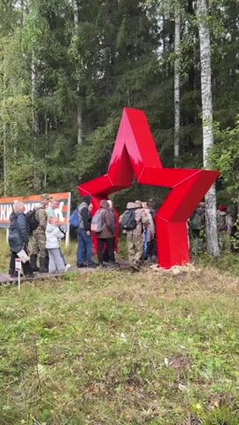 A group of people are gathered around a large, bright red, five-pointed star structure that is situated outdoors amongst trees. The star appears to be hollow, and some individuals are looking into it or interacting with it. The setting is a wooded area with grass and fallen leaves on the ground. There is a sign with orange and white lettering to the left of the star, but the text is partially obscured. The people are dressed in casual outdoor clothing, including jackets and backpacks, suggesting they are on an excursion or tour. The weather appears to be overcast or mild, as people are wearing layers.