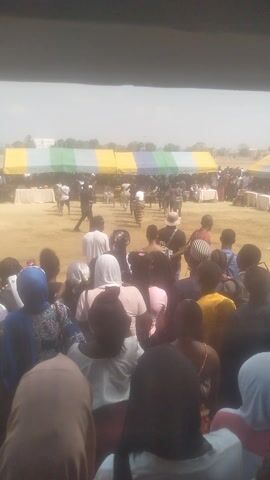 A crowd of people watches a performance taking place under a large tent. The tent has a colorful striped pattern and is set up on a sandy field. People in the crowd are wearing a variety of clothing and head coverings. Some individuals are standing, while others are seated. The performance appears to involve dancing, with several figures in the center of the field moving rhythmically. The atmosphere suggests a community event or celebration.