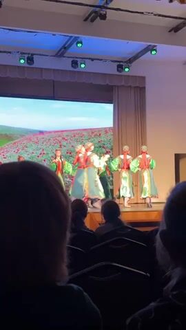 The video shows a group of dancers performing on a stage, viewed from the audience. The dancers, all wearing green and red costumes, are moving in a circle. Behind them is a large screen displaying a field of red flowers under a blue sky. The stage is well-lit with multiple lights, including those pointing down from the ceiling. An audience is seated in the foreground, with the backs of their heads visible. The room appears to be a theater or auditorium, with draped curtains on the side of the stage.