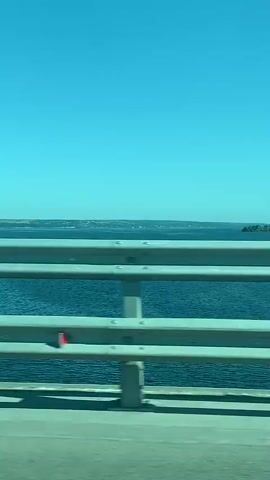 The video shows a view from a moving vehicle, likely a car, crossing a bridge over a body of water. The sky is clear and blue. A metal guardrail with multiple horizontal bars is visible in the foreground, separating the road from the water. The water is a deep blue and appears slightly choppy. In the distance, a shoreline is visible with some vegetation and structures. A small, green island or landmass can be seen in the water to the right of the frame. The road surface has a yellow line marking the edge of the lane. The perspective suggests the vehicle is traveling at a moderate speed.
