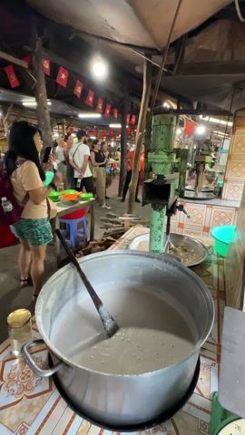 A tourist attraction in Vietnam features a large metal pot filled with a light gray liquid, being stirred with a wooden oar-like implement. In the background, tourists observe an individual in an orange shirt preparing small dishes. A green machine stands nearby, presumably involved in the process, and Vietnamese flags adorn the scene, enhancing its local character.