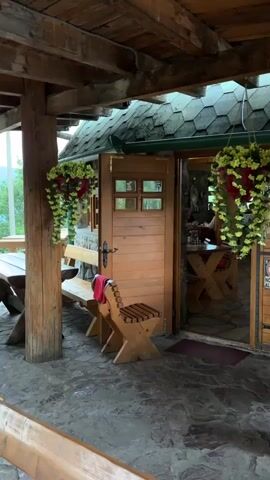 The video shows the exterior of a rustic restaurant or cafe, likely in a mountainous or rural setting given the wooden architecture and stone patio. The building features a wooden roof with shingles, exposed wooden beams, and large wooden support columns. In front of the entrance is a stone patio with several wooden picnic tables and benches. A single wooden chair with a red cloth draped over it sits near the entrance. Two large hanging baskets filled with yellow flowers and red accents are suspended from the overhang near the doorway.

The entrance itself is a double wooden door, one side of which is ajar, revealing the interior of the establishment. Through the open door, we can see more wooden tables and chairs, a stone floor, and a person sitting at one of the tables. The overall aesthetic suggests a cozy, traditional, and possibly historic establishment.

Visible text includes a sign near the right-side entrance that reads "NO DOGS ALLOWED". There is also some text in a foreign language, possibly Cyrillic, above the doorway on the right. The lighting suggests it is daytime, and the weather appears to be fair. The scene evokes a sense of tranquility and offers a glimpse into a charming, nature-inspired dining or resting spot.