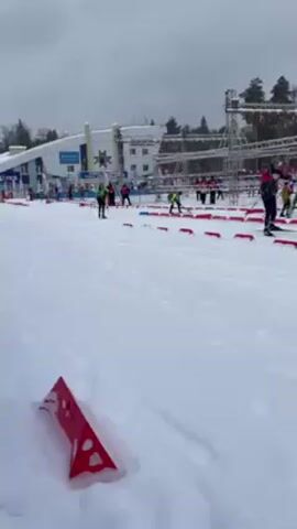 The video shows several people cross-country skiing across a snowy, cold environment. They are maneuvering around red and blue square blocks on the ground. Some of the skiers are wearing colorful outfits, while others wear green jackets. There are other people standing near the course watching the skiers. A tall metal structure is also visible in the background. Some tall buildings with signage are visible too. The ground is covered with tracks from skis.
