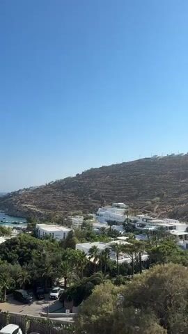 The video shows a panoramic view of a coastal town with white buildings and lush greenery under a clear blue sky. The scene overlooks a bay filled with numerous boats anchored in the turquoise water. In the foreground, there are some parked cars and vegetation. The landscape is characterized by a dry, rocky hillside sloping down towards the sea.