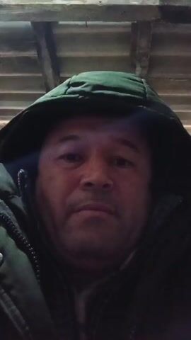 The video shows a close-up shot of a man's face. He is wearing a dark green hooded jacket. The hood is up, partially obscuring his forehead. His face is somewhat weathered, and he has a serious expression. The background appears to be the underside of a wooden structure. The lighting is dim. He inhales audibly at one point.